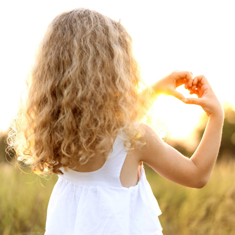 Mädchen mit blonden, lockigen Haare das mit ihren Händen ein Herz formt